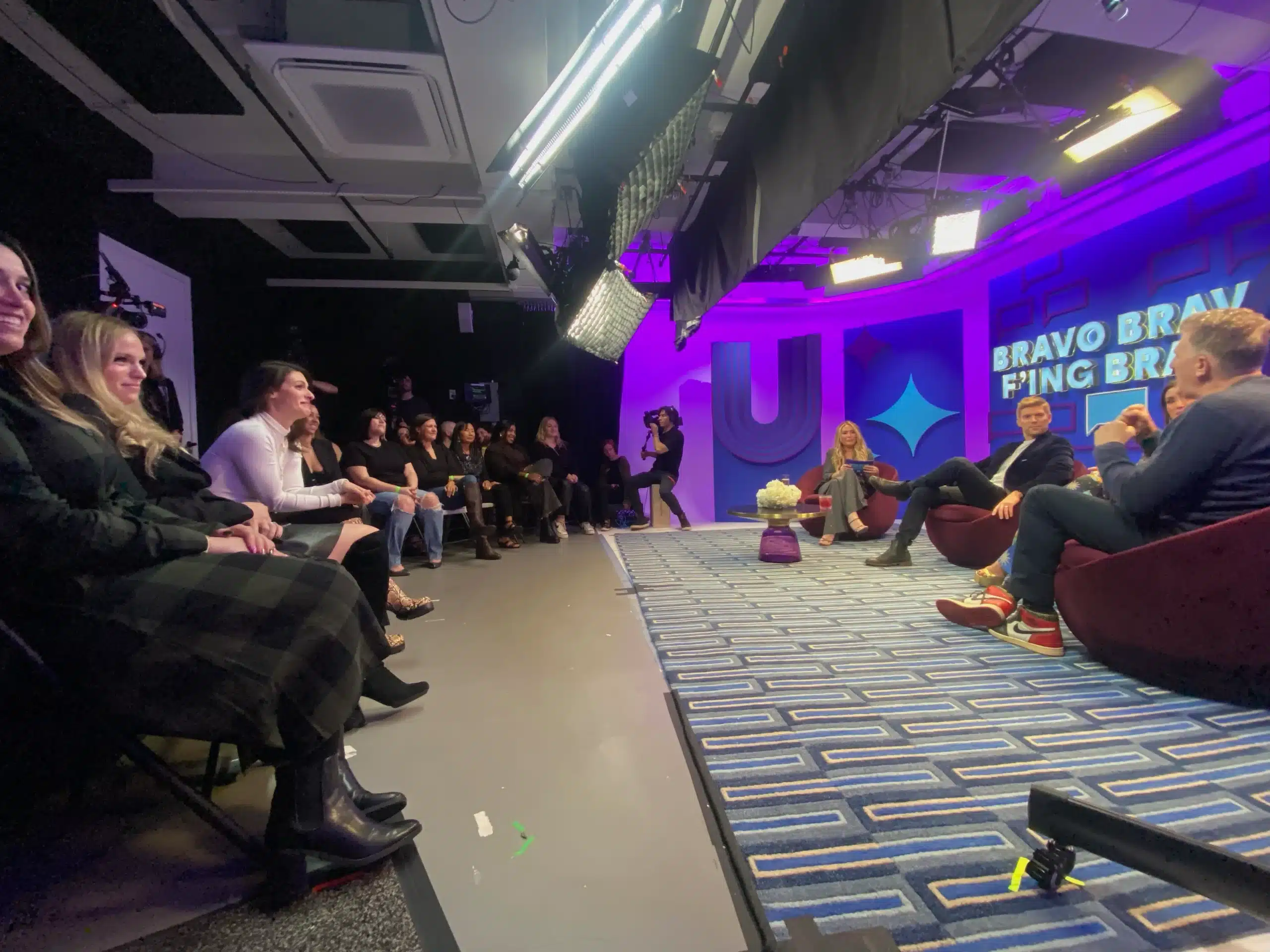 A panel discussion is taking place in a brightly lit studio with purple and blue lighting, set up for video production. Four people sit on stage talking, while an audience watches and listens attentively. A sign reads “BRAVO BRAV FING BRAVO.”.