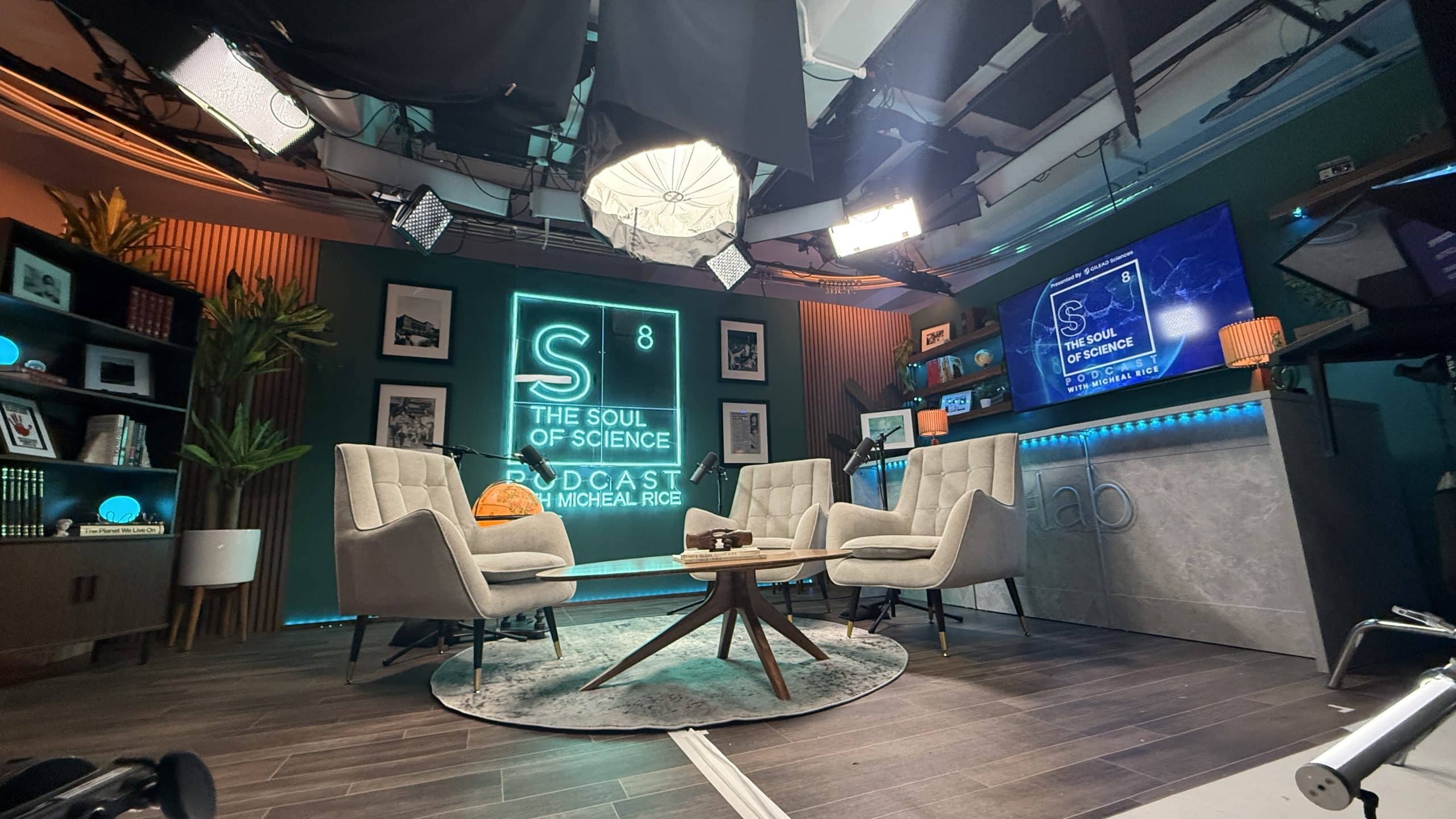 A modern podcast studio with two beige chairs, a small round table, studio lights, and a neon sign reading “S8 The Soul of Science Podcast with Michael Roe.” Bookshelves and monitors fill the stylish, well-lit room.