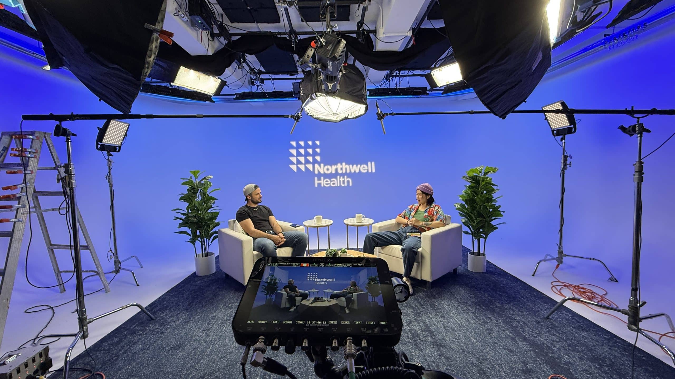 Two people sit in armchairs on a set with a blue background, “Northwell Health” logo behind them. Camera equipment, lights, and cables are visible, with a camera monitor in the foreground. Potted plants are beside each chair.