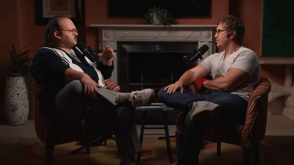 Two men, reminiscent of Stavros Halkias and Matt Johnson, sit across from each other in armchairs, engaged in conversation with microphones in hand, in a warmly lit living room setting with a fireplace in the background—a scene that could easily unfold on the A24 Field Podcast.