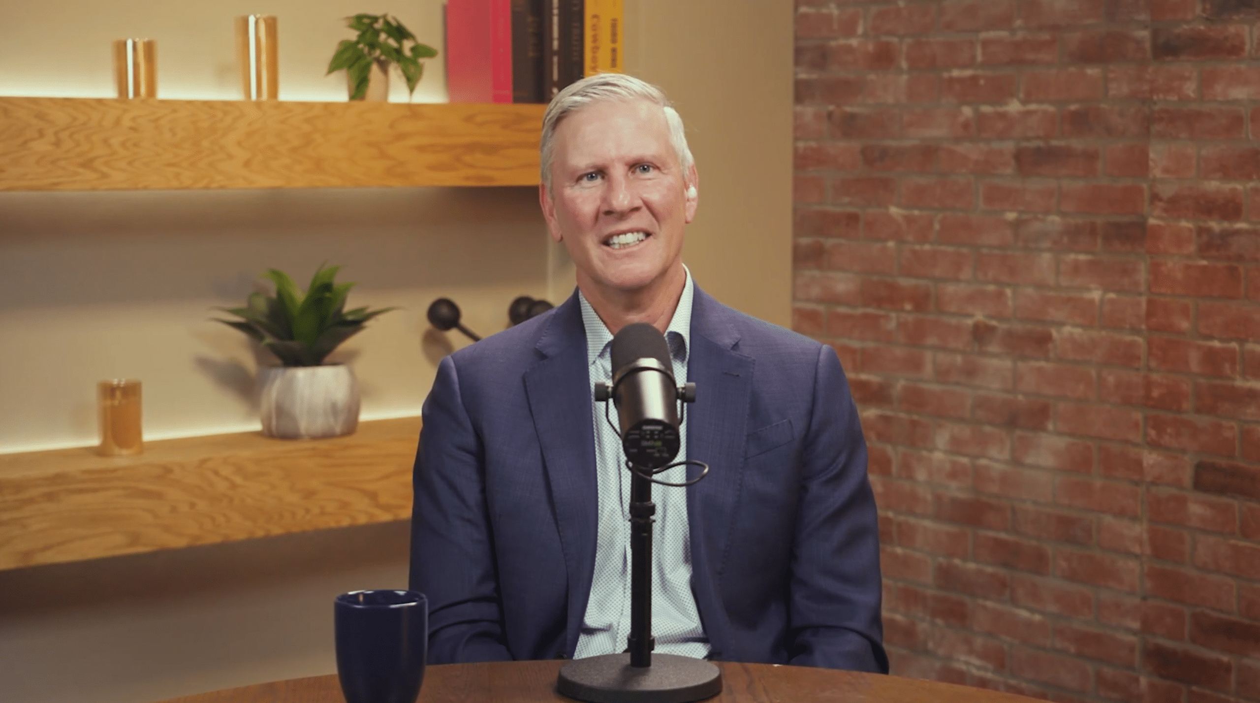 A man in a blue suit sits at a table with a microphone, smiling, as if hosting a show at ASL Studios. There is a blue mug on the table and shelves with plants and books in the background, evoking a professional yet inviting productions setting. A brick wall is partially visible.