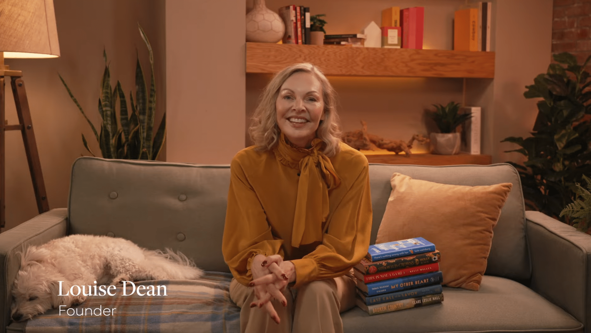 A woman sits on a sofa next to a stack of books, smiling at the camera inside a cozy Video Studio NYC. A white dog lies on the sofa. Warm lighting and bookshelf decor are in the background. Text reads: “Louise Dean Founder.”.
