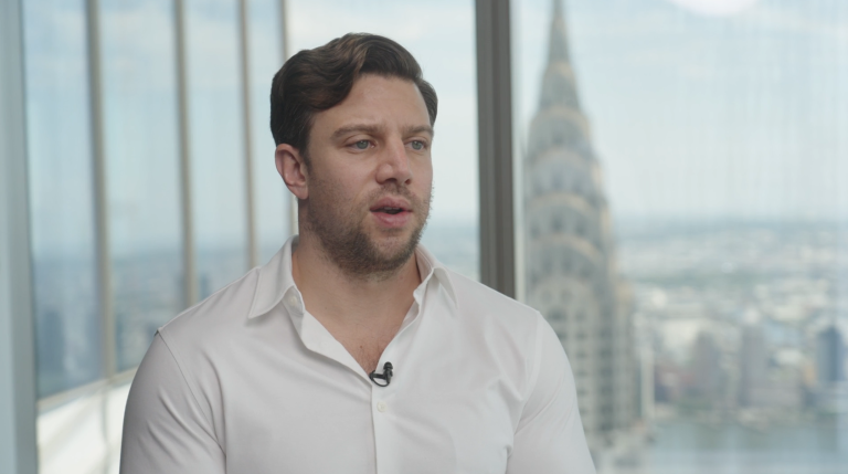 A man in a white shirt speaks in front of a large window with a cityscape and the Chrysler Building visible in the background, creating an impressive setting for high-quality video production.