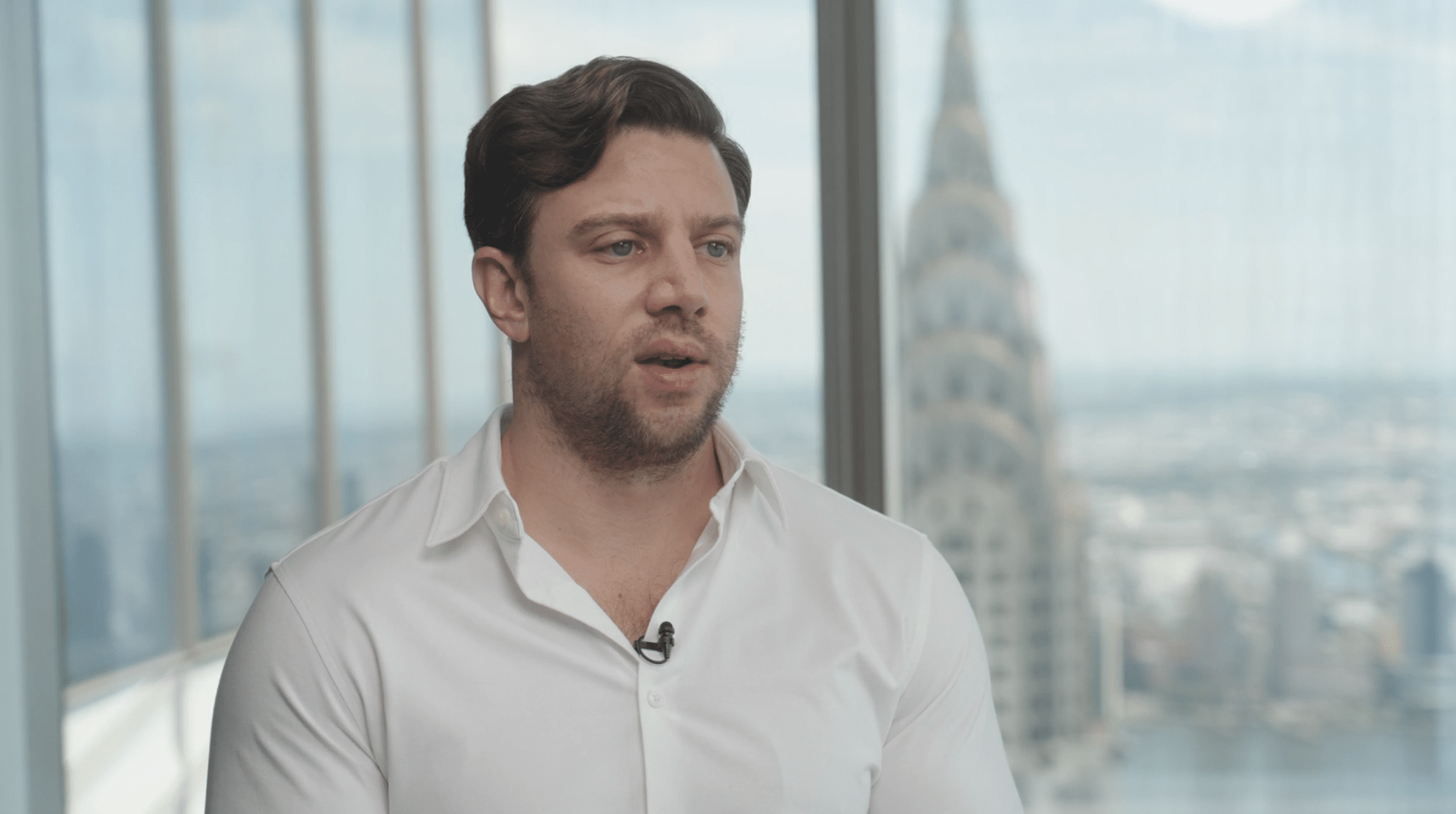 A man in a white shirt speaks in front of a large window with a cityscape and the Chrysler Building visible in the background, creating an impressive setting for high-quality video production.